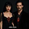 A woman and a man in black attire wearing ornate red, black, and gold pendants, against a dark background with branding text at the bottom.